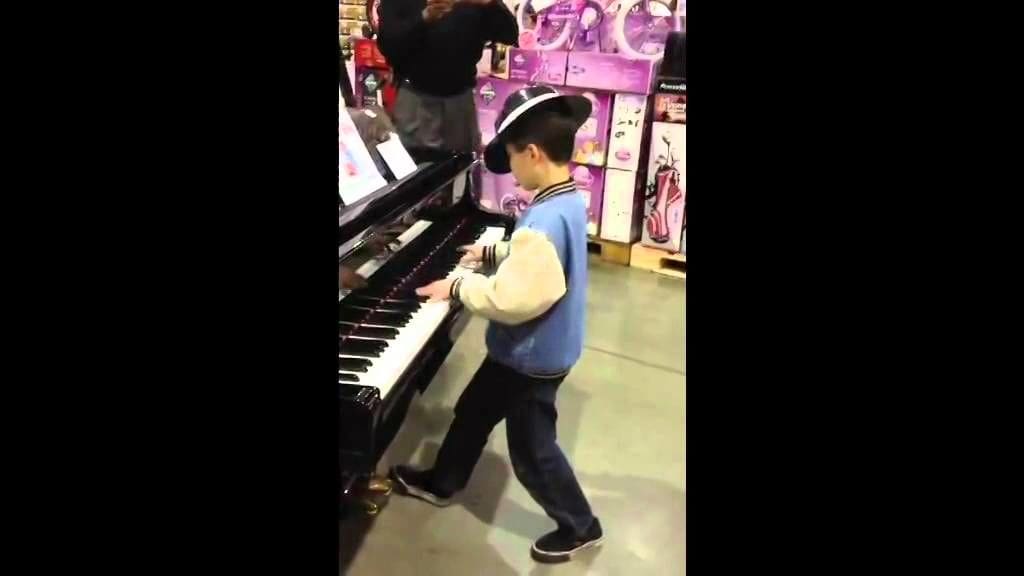 A child wearing a blue and white sweater with black trim plays the piano in what appears to be a toy store. Shelves stocked with various items, including pink packaged goods, are visible in the background.