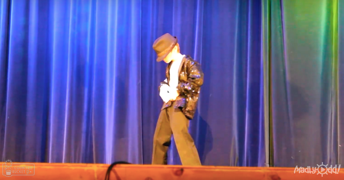 A male performer in a theatrical setting with blue curtains as the backdrop, wearing a loose-fitting brown jacket and beige pants on stage.