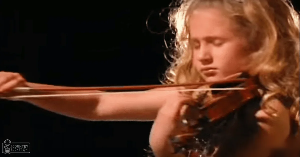 A young girl playing the violin indoors, dressed in dark clothing with warm lighting highlighting her and the instrument.