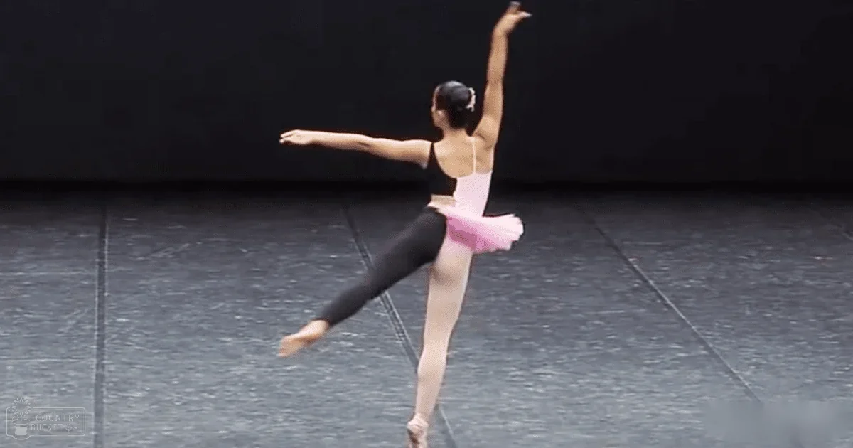 A young adult female ballet dancer in a studio, wearing a black leotard with a white undershirt, pink tutu skirt, and pointe shoes, styled up hair with an accessory.