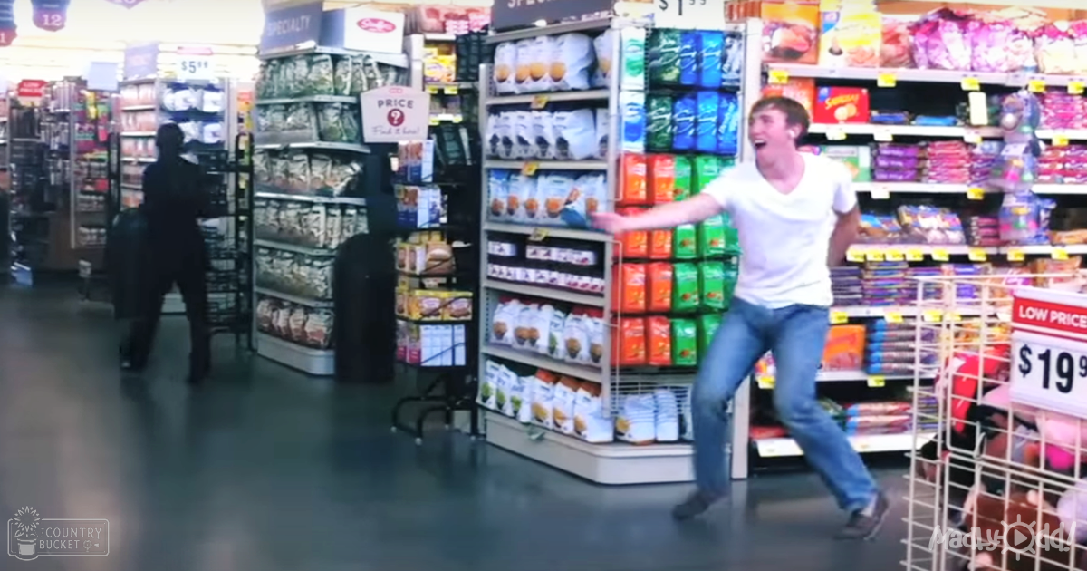 A young adult male in a grocery store aisle, wearing dark pants and a dark jacket or shirt with another person nearby laughing or shouting.