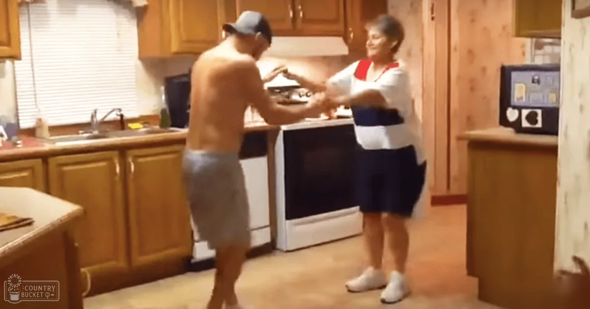 An older adult male singer in gray shorts, a dark cap, interacting with an older adult female wearing red-accented white polo and navy blue athletic shorts in a kitchen setting with natural light filtering through blinds.