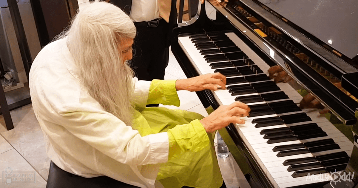 An older adult female in a white jacket over a lime green garment, playing a black piano indoors on a tiled floor in what looks like a public space during the daytime.