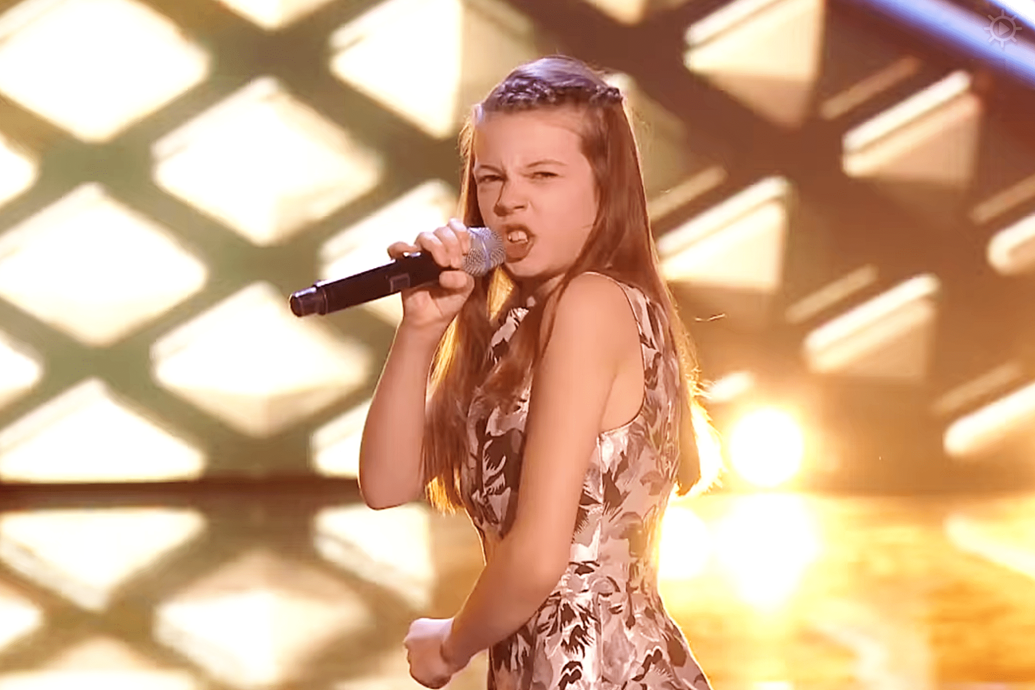 A young girl in a floral sleeveless dress performs on stage under warm, golden lights.