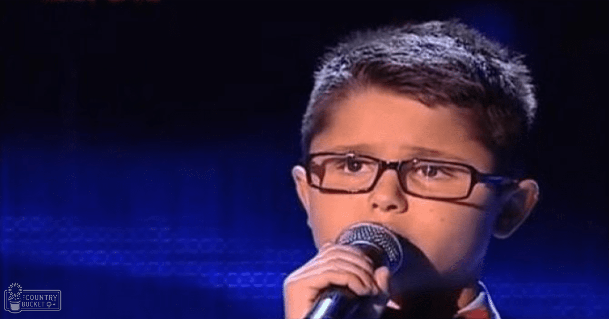 A young boy wearing glasses sings into a microphone at what appears to be a talent show setting with blue stage lighting.