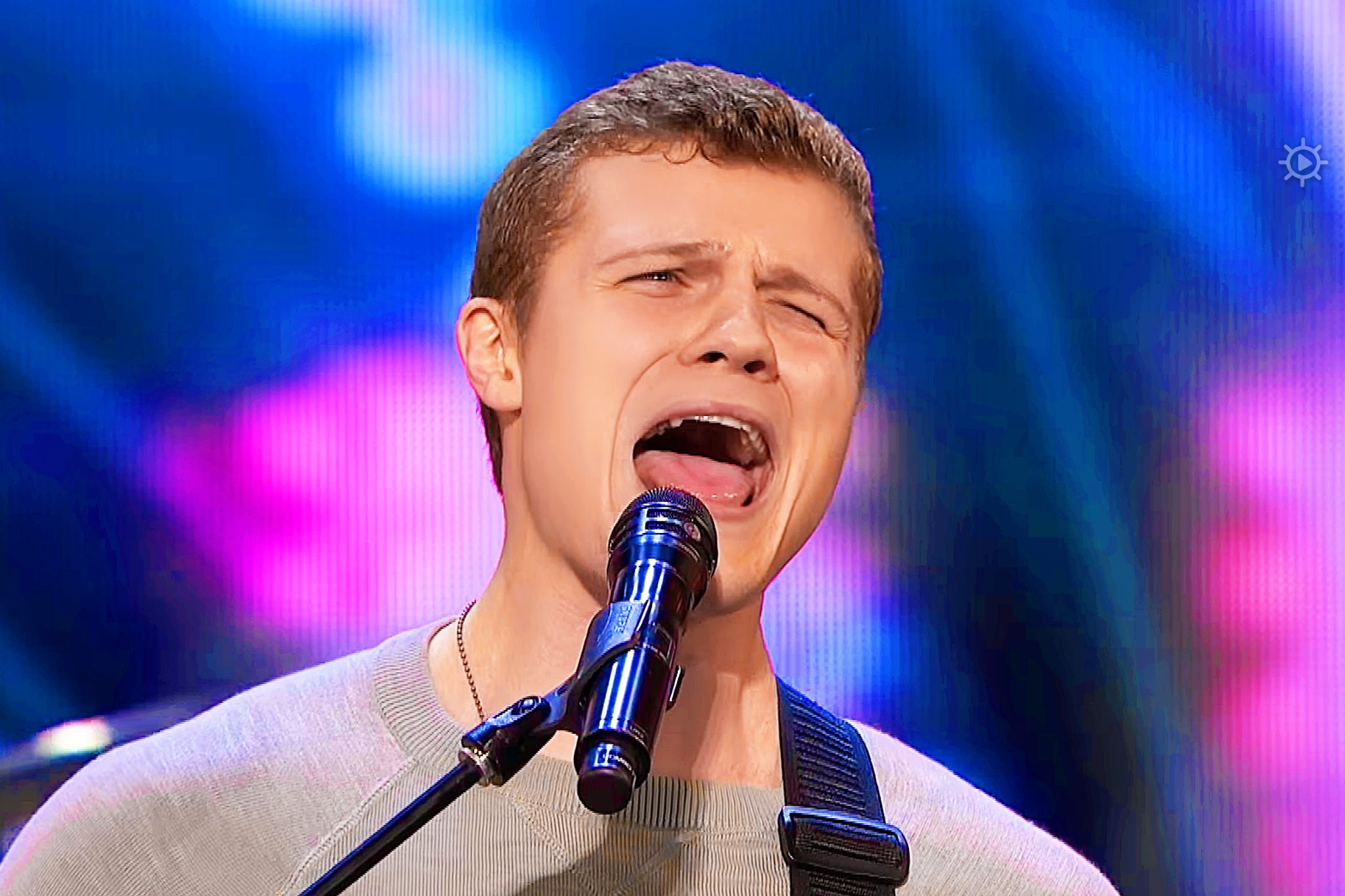 A young adult male singer wearing a light gray long-sleeved shirt and holding a microphone with a blue windscreen on stage, set against vibrant indoor performance lighting.
