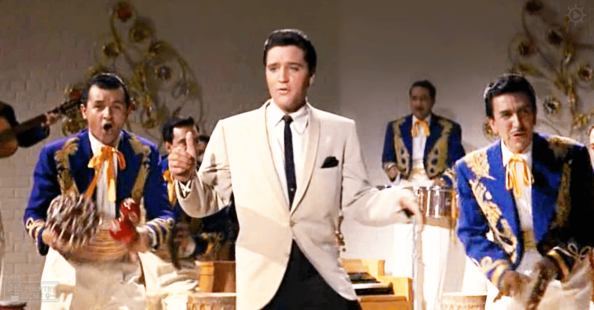 Adult male in beige suit jacket with black tie, surrounded by young adult musicians wearing blue jackets with gold trim and orange scarves. Indoor venue with musical instruments and ornate decorations.