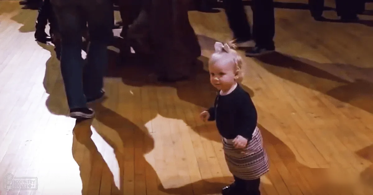 A toddler with a ponytail and bow, wearing a dark long-sleeved top and striped skirt, indoors in a public space during daytime with brown wooden flooring.