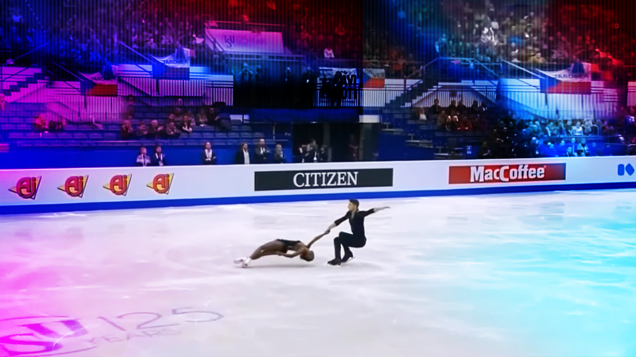 A young adult female figure skater in a black leotard and white figure skates performs on an indoor ice rink with tiered seating filled by spectators dressed in formal attire, under evening or night artificial lighting. Advertisements for 'CITIZEN' and other sponsors are visible around the rink.
