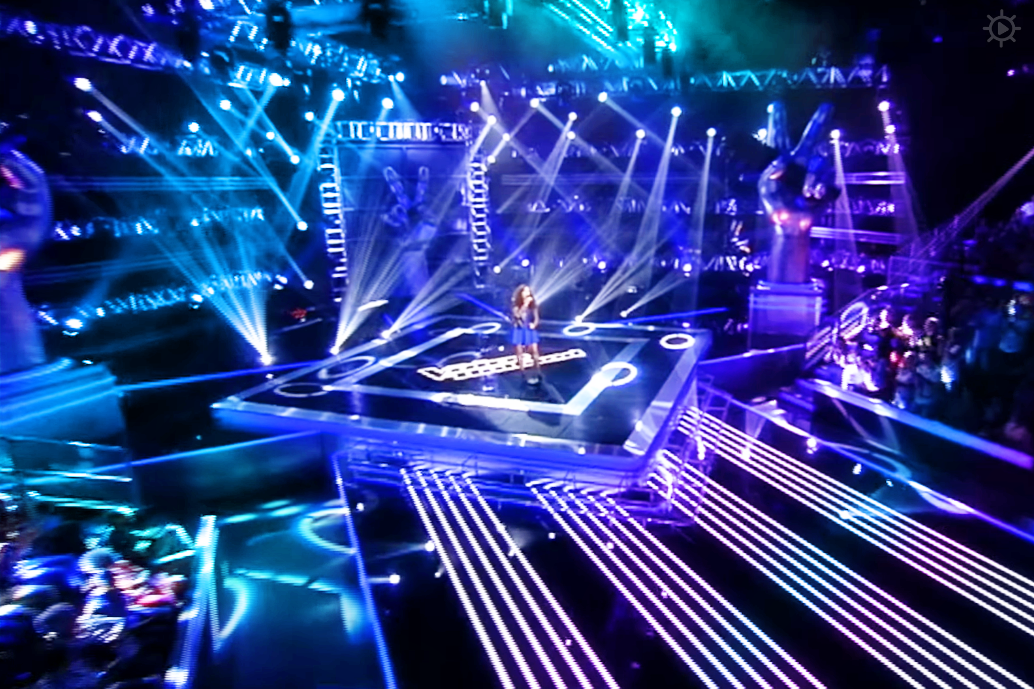 A female adult singer in a blue sleeveless short dress performs on stage under elaborate lighting at an indoor concert or show venue.