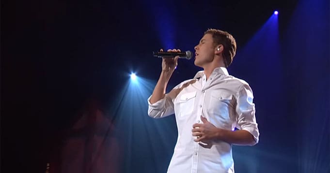 A young adult male in a relaxed white button-up shirt performing on a stage with spotlights, likely at an indoor concert hall or theater.