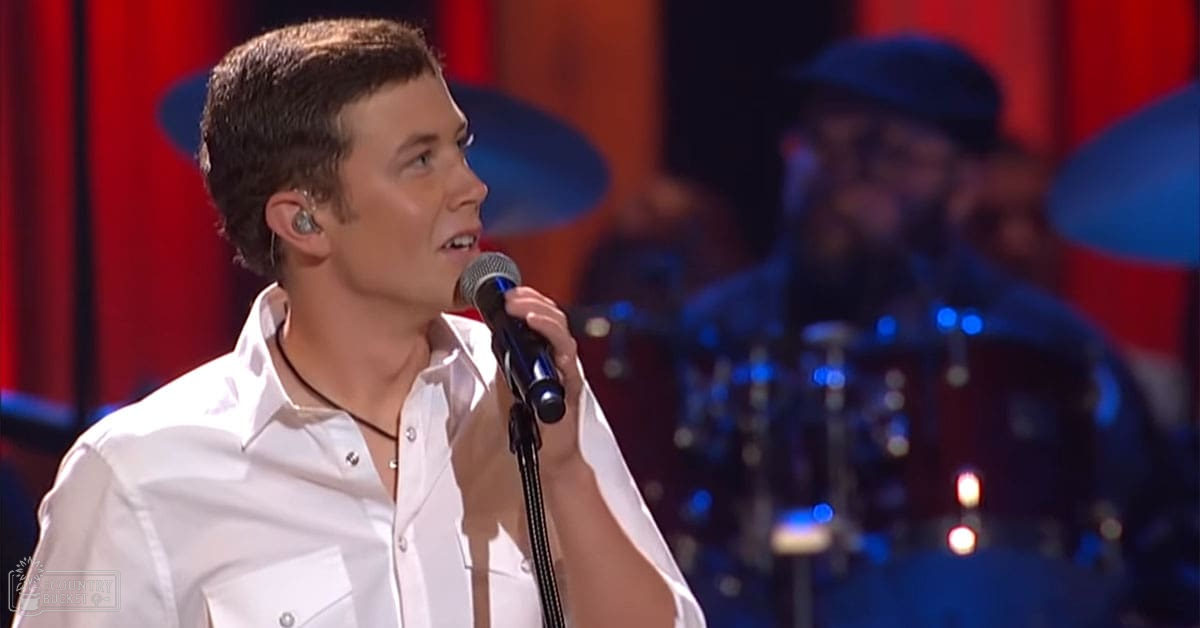 A young adult male singer wearing a white button-up shirt with an earpiece in one ear, performing in what appears to be a studio setting.