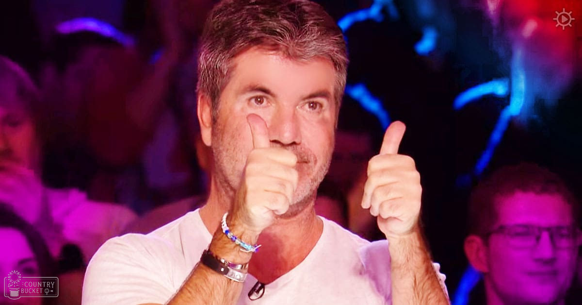A middle-aged man wearing a white t-shirt and bracelets in an indoor setting, possibly a television studio with stage lighting and audience members behind him.