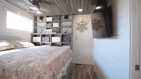 A cozy bedroom with a bed, shelves holding books and decor, ship's wheel wall decorations, and ceiling fan in natural light.