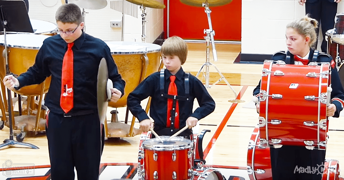 Children playing drums inside a school gymnasium with wooden floors and white-red walls, wearing black shirts with red ties and black pants; drumsticks visible.