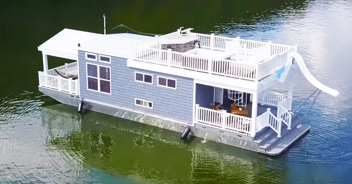 A serene houseboat with blue siding, white trim, and a slide in calm water, surrounded by an expansive outdoor setting.