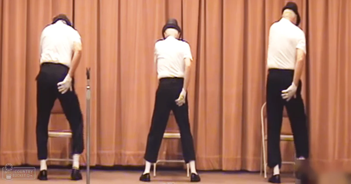 Middle-aged adult males performing a dance routine in an indoor community setting with simple chairs and beige curtains, bright lighting suggesting daytime or artificial illumination.