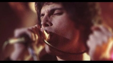 A young adult male in a tailored tuxedo jacket and white shirt plays a trumpet on an indoor stage, illuminated by warm evening lighting.