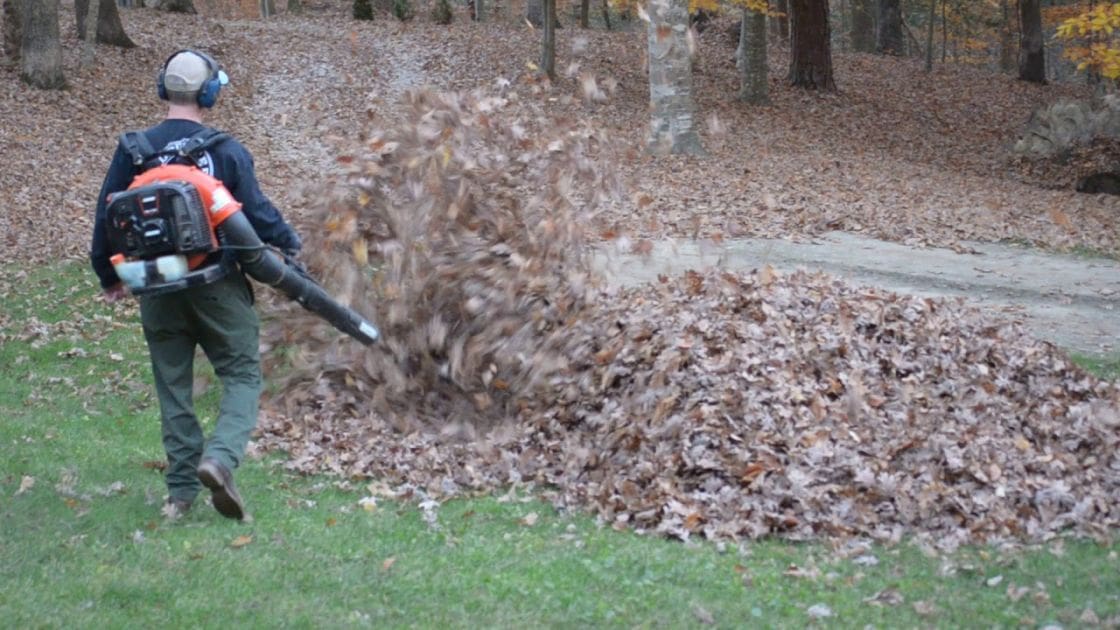 An adult male wearing a dark-colored long-sleeved shirt with a logo or design on the back, using a backpack-style leaf blower in an outdoor yard setting during autumn.
