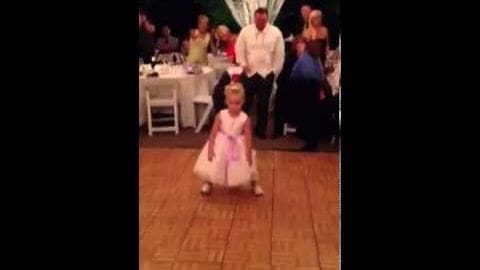 A young child in a white dress with pink accents stands formally, wearing ballet slippers. Adults are seated behind the child at a table covered by a white cloth, suggesting an indoor social event.