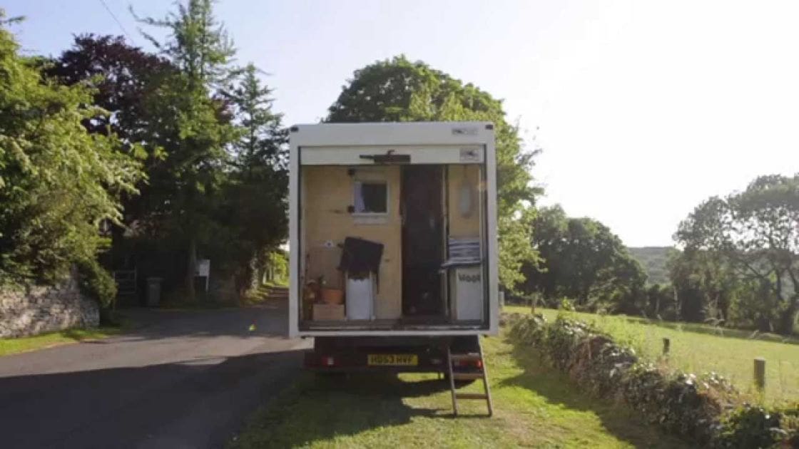 A white mobile home with open door and side panel, surrounded by lush greenery, on a paved rural road under bright daylight.