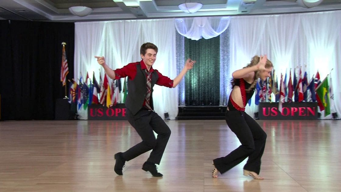 A dance duo performing at an indoor ballroom event called 'US Open,' with a teenage male dancer wearing red and black attire, and a female dancer in a black outfit with red accents, barefoot. Flags from various countries are displayed behind them.