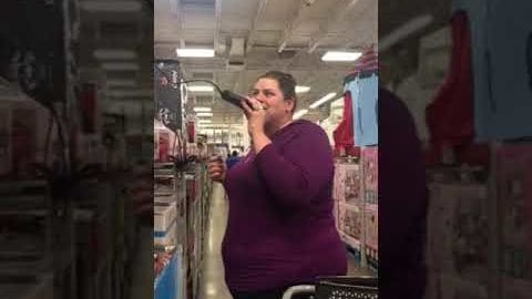 Middle-aged female holding a microphone in an indoor retail setting with purple long-sleeve shirt and shopping cart handle visible.