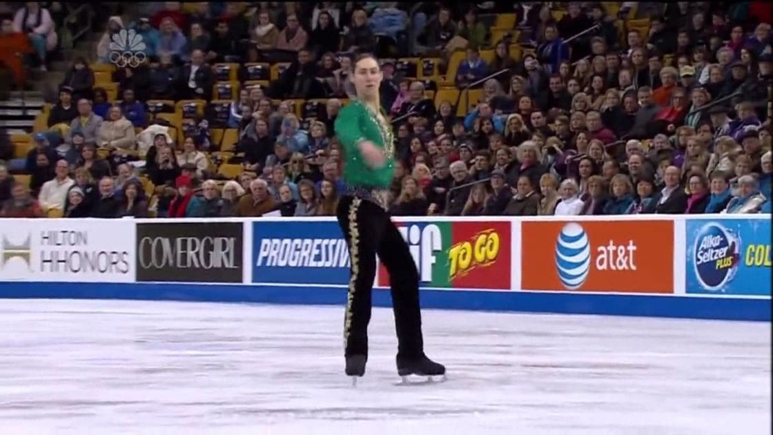 A young adult male figure skater performing at an indoor ice rink filled with spectators, illuminated by artificial lights typical of a sports venue.