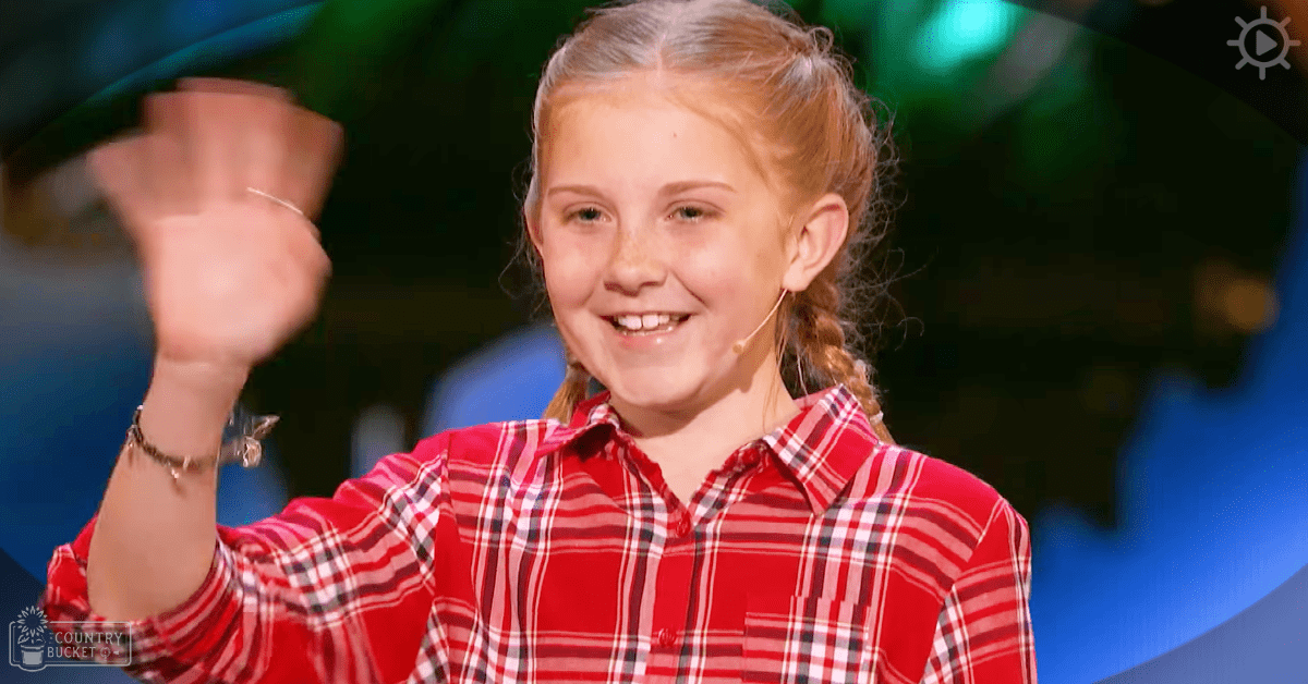 A young girl wearing a red plaid shirt with white and black stripes stands indoors, likely at an event or performance venue given the stage-like setting.