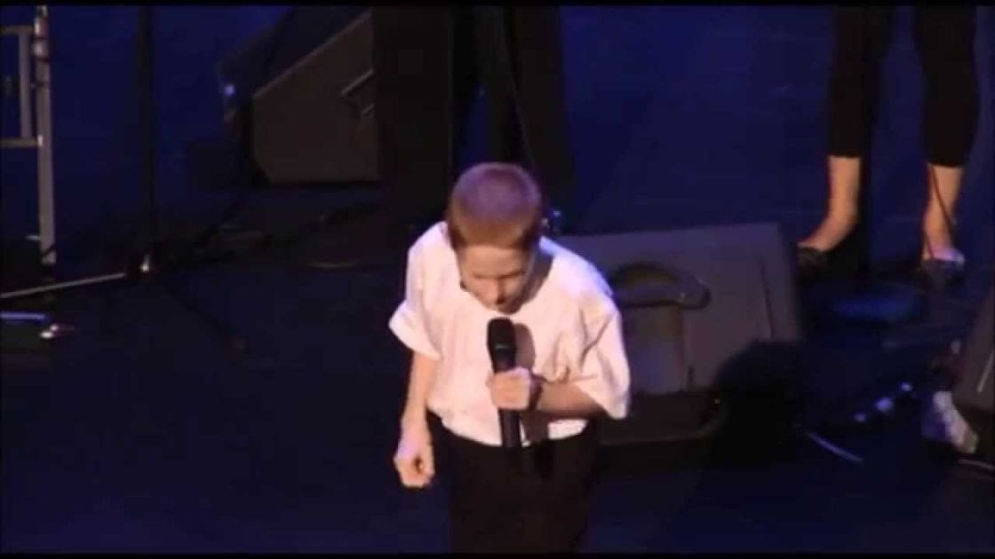 Young adult male singer performing on a dark stage with microphones and speakers in an indoor venue during evening lighting.