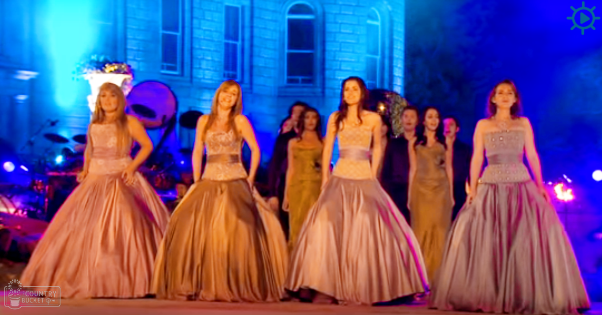 A group of young adult women in elegant dresses at an outdoor prom-like event with artificial lighting and a building in the background.