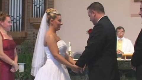 A bride in a white strapless wedding gown with a full skirt and long veil stands next to her groom in a dark suit jacket at an outdoor wedding ceremony. They are surrounded by other guests, including a young woman in a maroon dress smiling nearby.