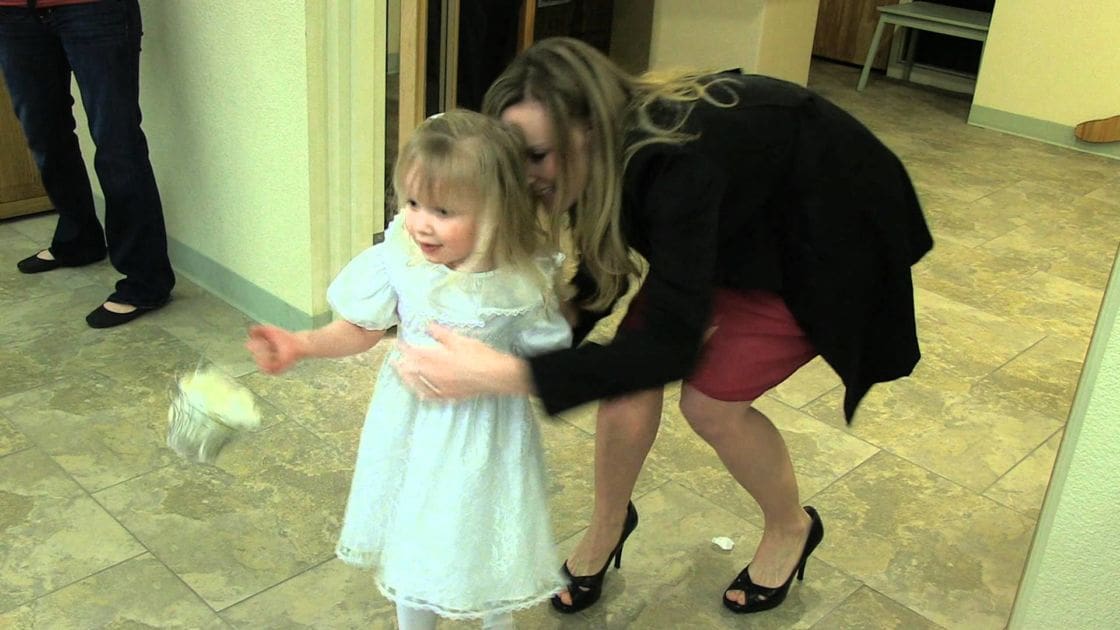 A toddler girl wearing a white lace-detailed dress indoors, accompanied by an adult woman in a black blazer and red skirt.