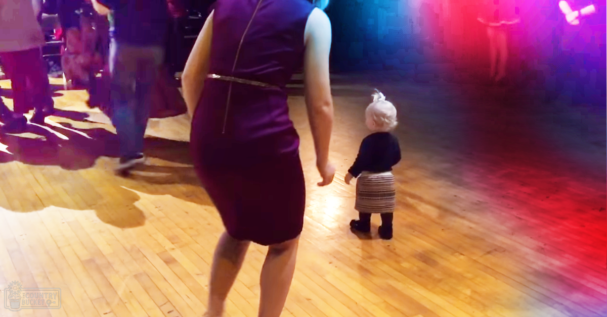 A toddler wearing a black long-sleeve shirt with horizontal stripes, striped skirt or pants, dark boots, dancing on a wooden dance floor in an indoor venue under red and blue lighting at night; accompanied by an adult female in a purple dress with a metallic belt, possibly also dancing.