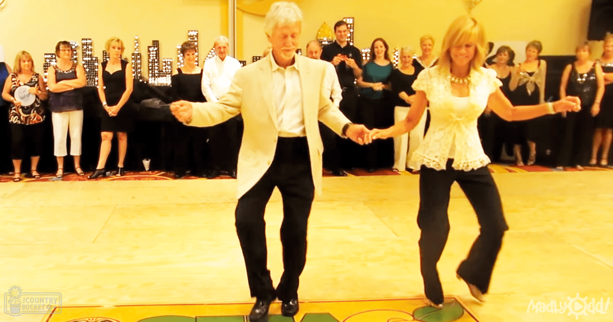 A couple dancing indoors at a social gathering, dressed formally under artificial lighting with decorative wall lights.