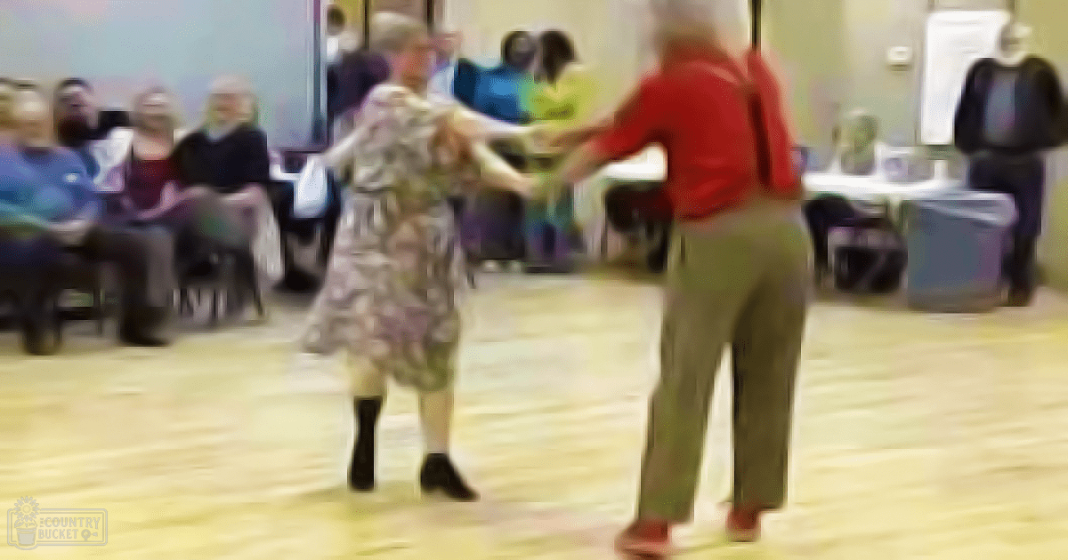 An older adult couple dancing in a community center with spectators seated around, wearing red and floral attire.