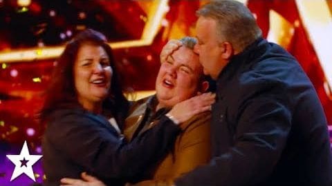 Adult female in dark blazer smiles at middle-aged man embracing adult male from behind on a vibrant red and gold stage with star logo, likely during an evening talent show event.
