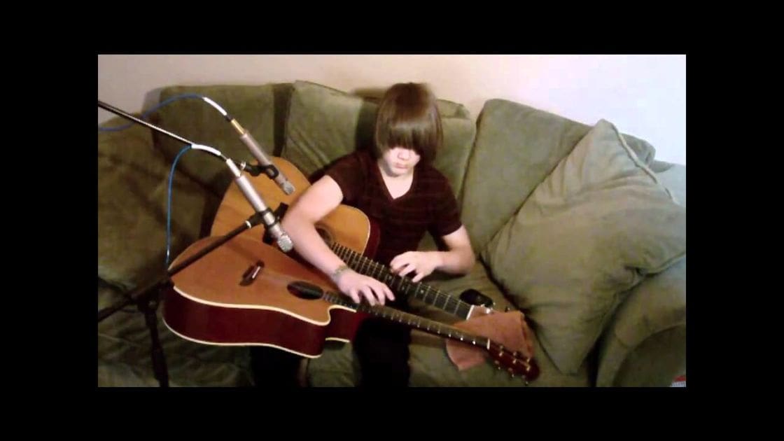 A young adult female playing guitar in a casual living room with green furniture and minimal decor, suggesting an evening ambiance.