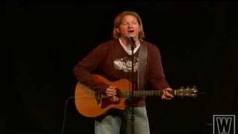 A middle-aged male singer wearing a brown sweater over a white shirt with text and light-colored jeans, holding an acoustic guitar on stage in front of a microphone stand.