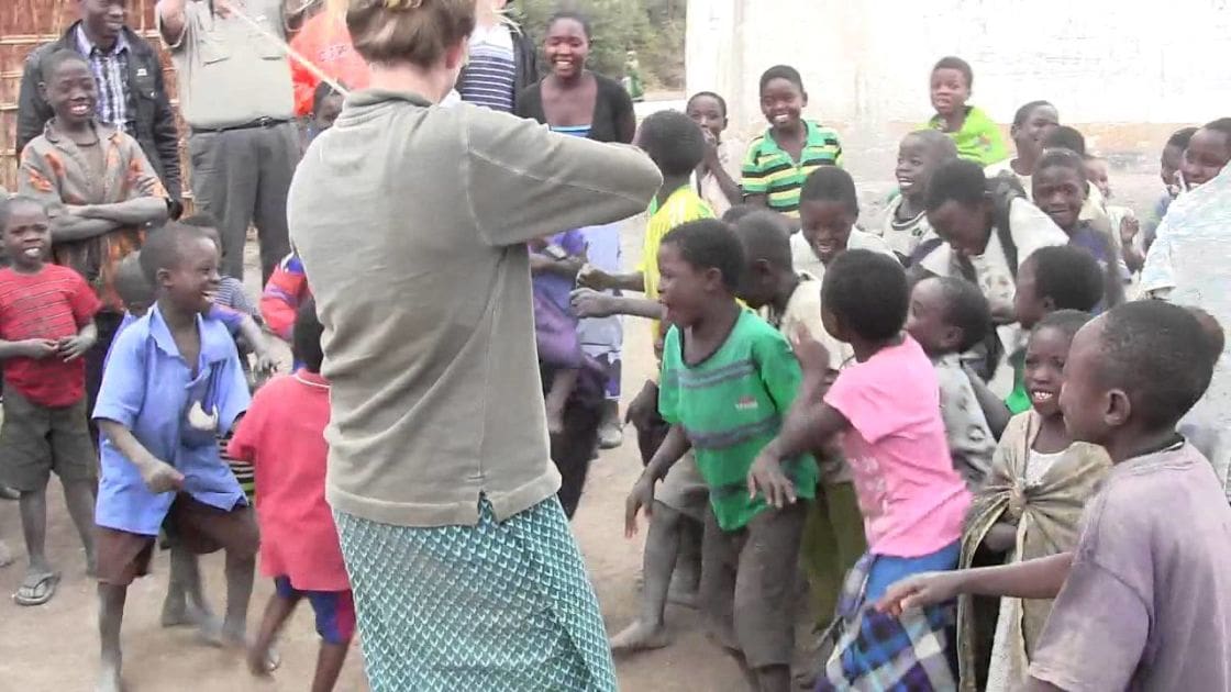 A child in a light blue shirt and sandals stands outdoors among adults and other children on a dirt ground with simple rural buildings in the background.