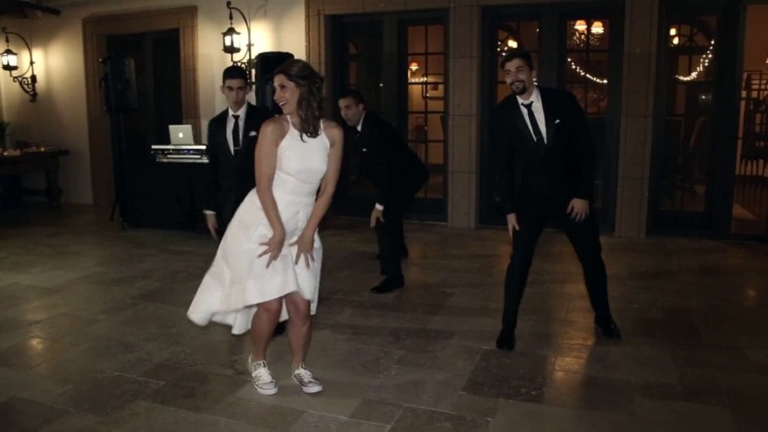 A woman in a white sleeveless dress with a high-low hemline dances alongside two men in black suits at an indoor formal event, possibly a wedding reception. String lights and DJ setup indicate evening ambiance.