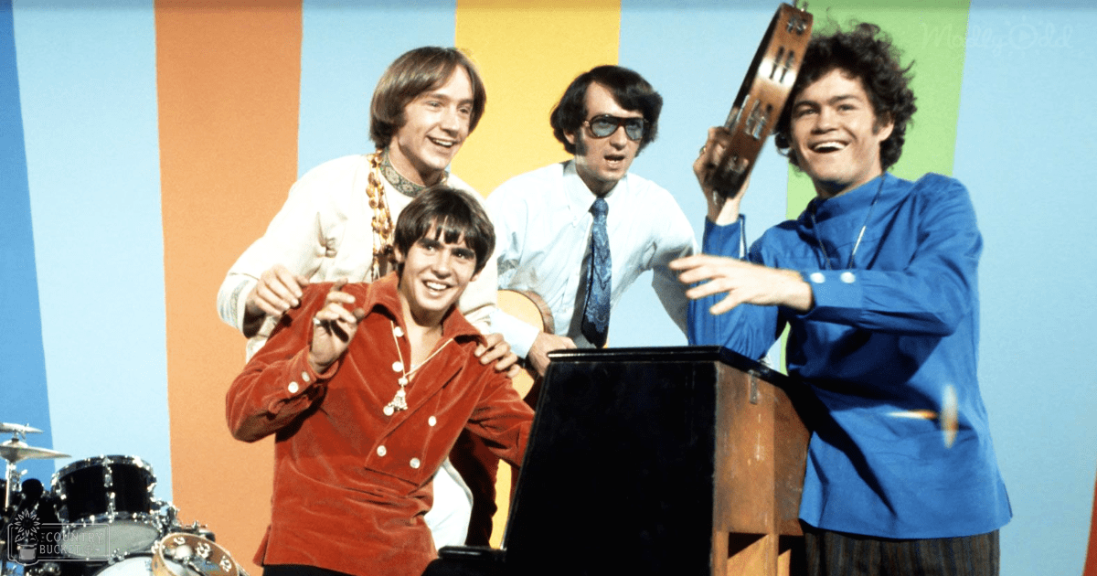 A musical group performing on stage with a colorful backdrop; young adult male singer wearing a red velvet jacket with white buttons and gold chain necklace in foreground left.