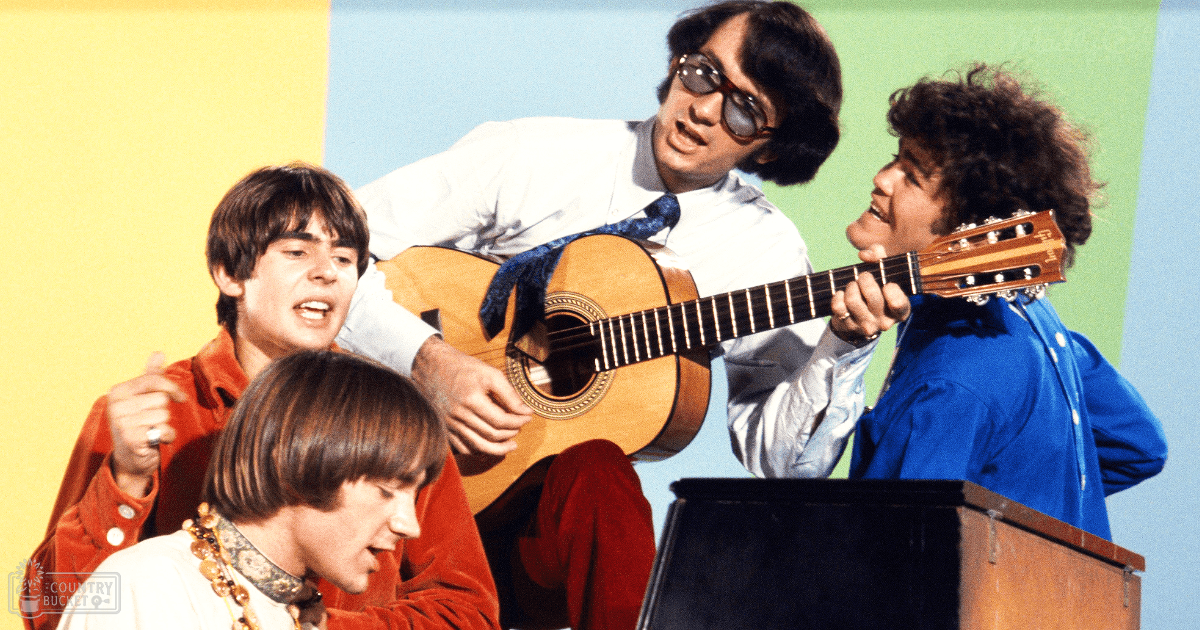 A musical performance scene from the 1960s with four young adult males in a stage setting, wearing formal attire and holding guitars against a colorful backdrop.