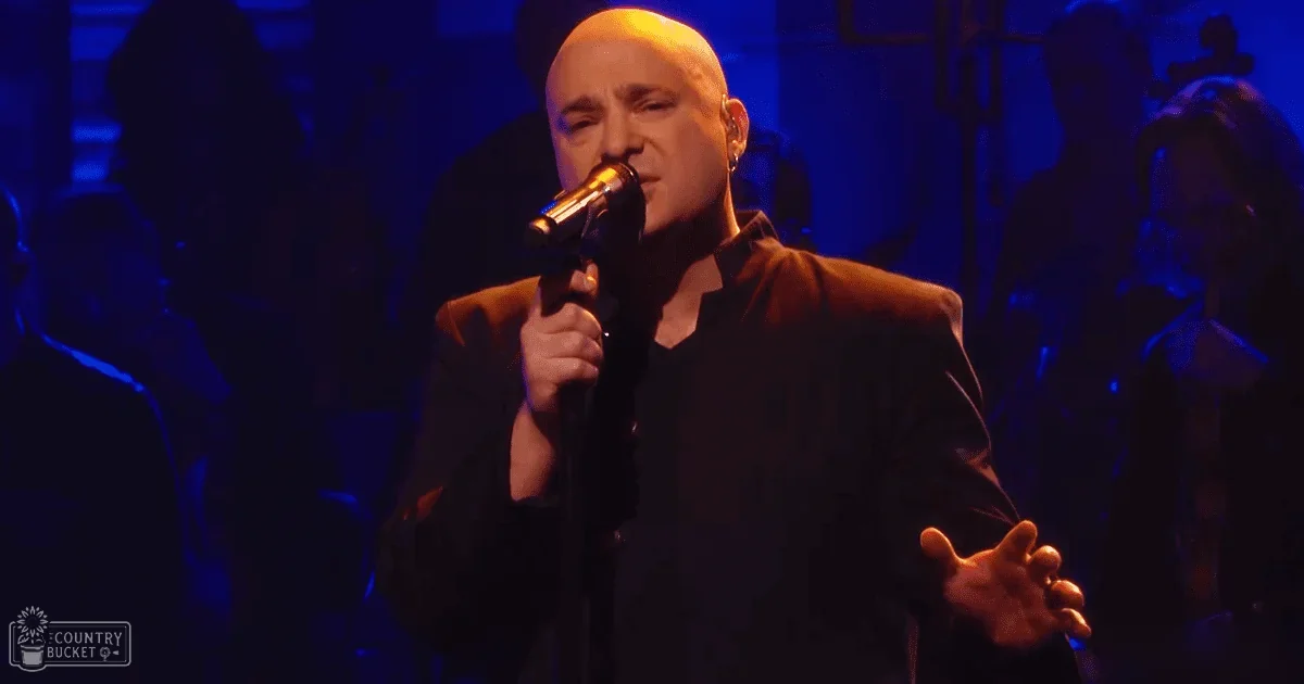 A middle-aged male singer wearing a dark jacket and V-neck shirt performs into a microphone at a dimly lit concert venue.