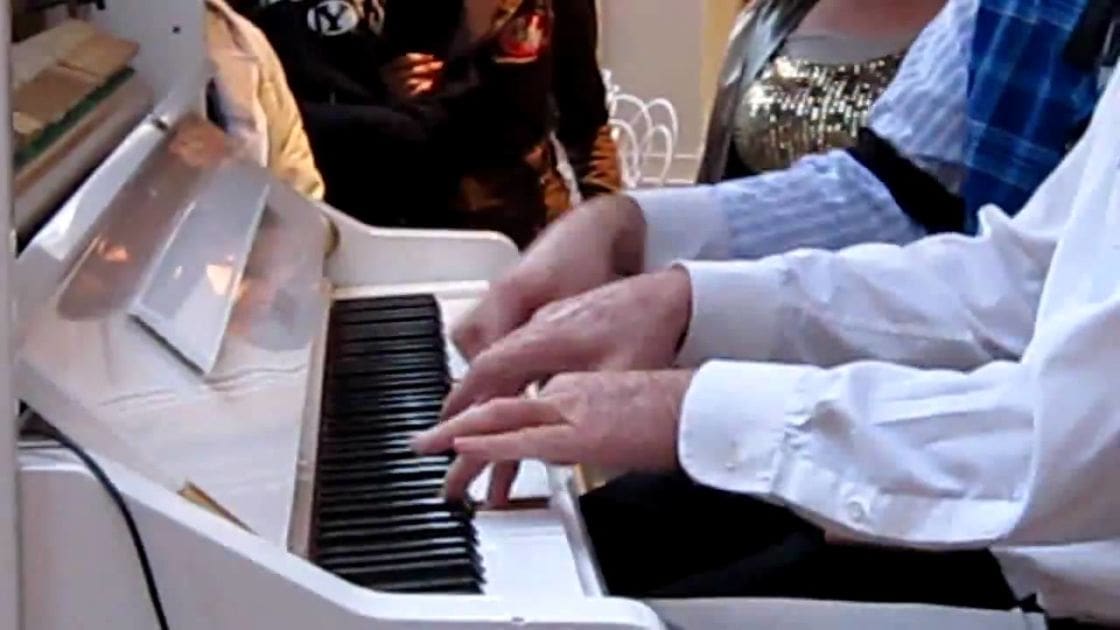 An older adult male pianist plays an upright piano in a public setting with natural light, surrounded by other people and objects.