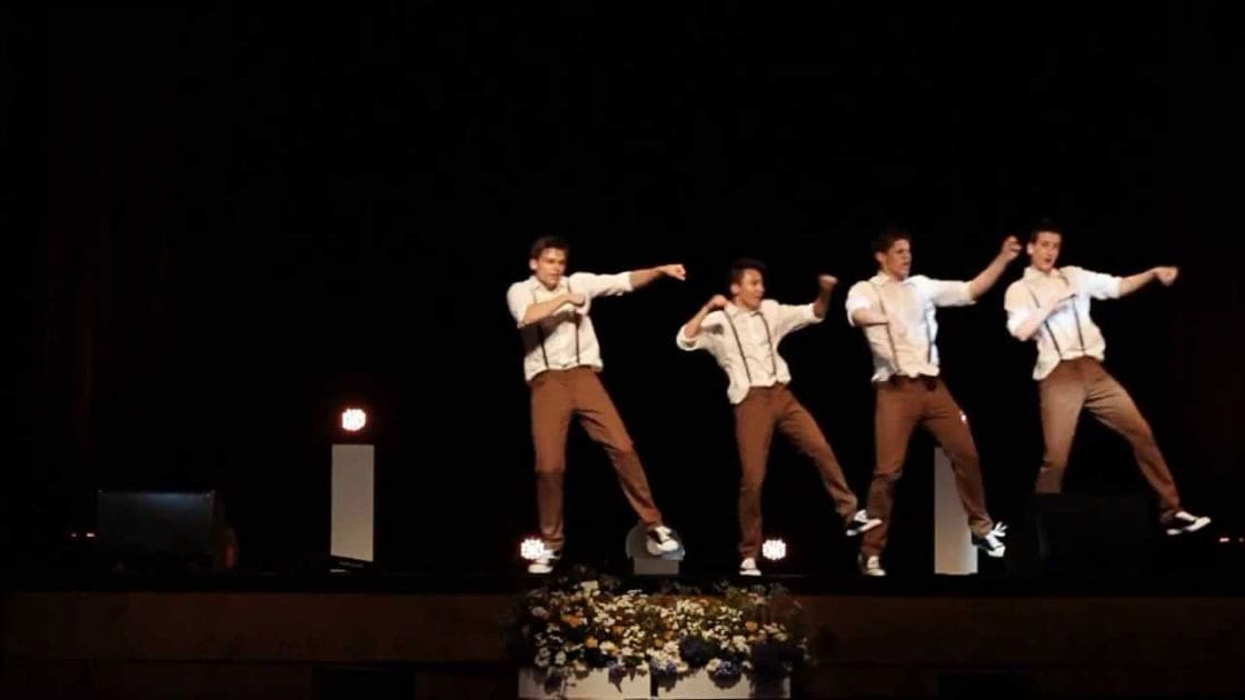 A group of young adult male dancers in a 1940s-style swing dance outfit perform on stage, with dimly lit background and floral arrangement at the front.