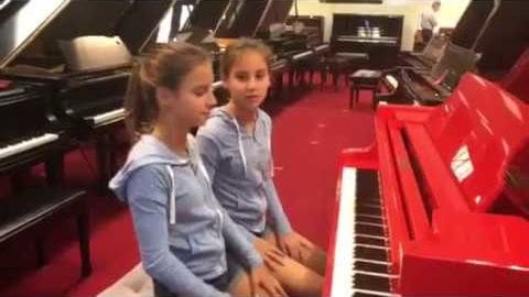 Two children in a music studio, wearing casual hoodies, surrounded by grand pianos with red carpet flooring.