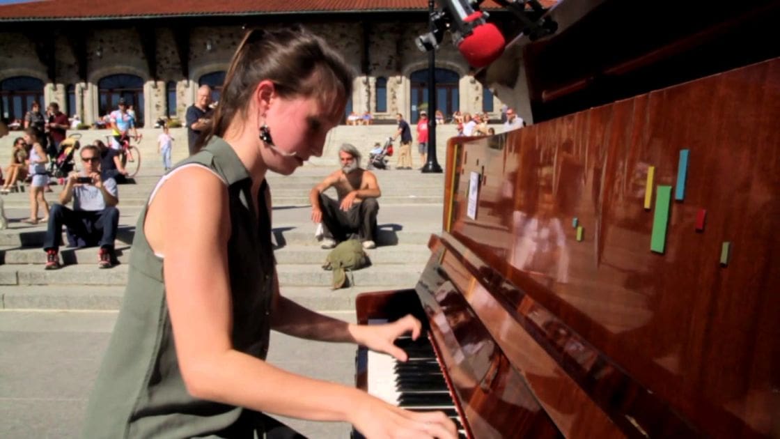 A young adult woman plays piano outdoors on stone steps with others casually gathered around, under bright daylight.
