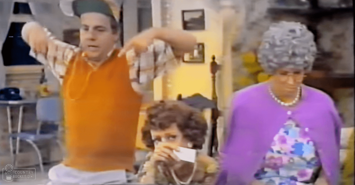A young adult male wearing an orange plaid shirt indoors with other adults, a child, furniture, and wall decor under natural light.