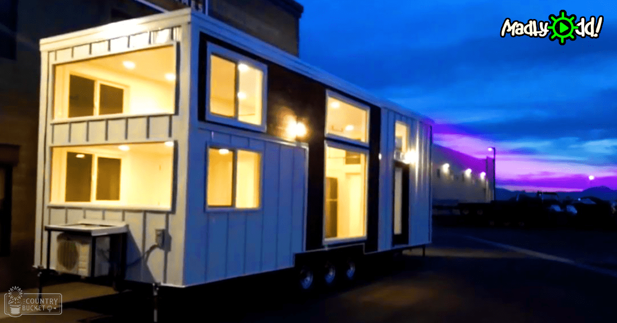 A two-story modern tiny house on wheels with large illuminated windows set in an urban parking lot area under a twilight sky.